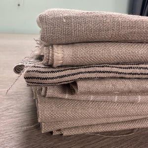 May include: A stack of folded fabric pieces featuring a coarse, woven texture in neutral beige tones. One piece of fabric in the middle of the stack displays a pattern of thin, horizontal black stripes against the light tan background.