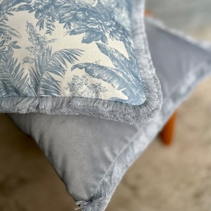 Könnte beinhalten: Ein quadratisches Zierkissen mit einem blau-weißen botanischen Toile-Muster auf der Vorderseite und einem einfarbigen, samtartigen grauen Stoff auf der Rückseite. Die Kanten des Kissens sind mit einer passenden grauen Fransenborte versehen.
