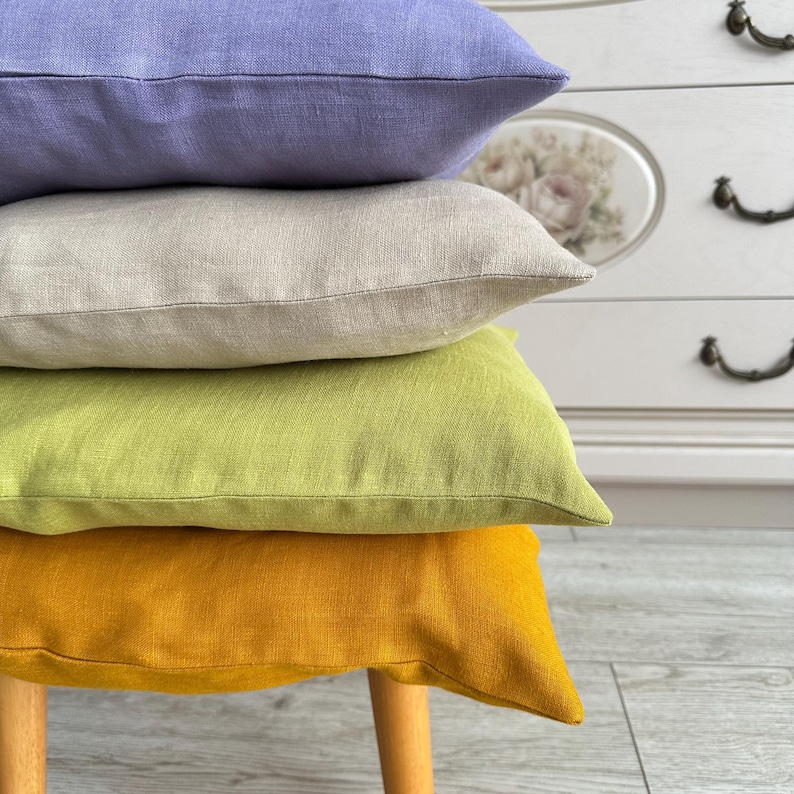 May include: A stack of four square throw pillows with a visible linen-like texture. The top pillow is lavender, the second is beige, the third is lime green, and the bottom pillow is mustard yellow. Each pillow has a simple, clean-edged design.