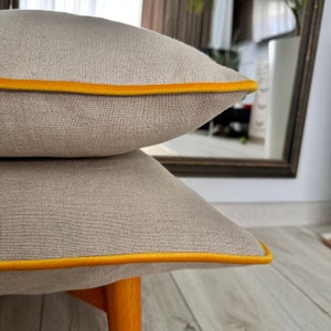 Peut inclure: Deux coussins de canapé en lin beige avec une bordure en velours jaune. Les coussins sont empilés l'un sur l'autre.