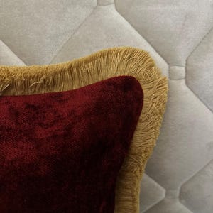 Pode incluir: Close-up de uma almofada decorativa com um centro de veludo vermelho escuro e uma borda com franjas douradas. A almofada está sobre um fundo acolchoado branco sujo, criando um contraste de texturas e cores. O design sugere um item de decoração de casa luxuoso.