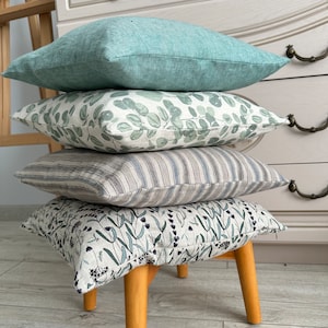 Könnte beinhalten: Ein Stapel von vier dekorativen Kissen mit verschiedenen Mustern. Das oberste Kissen ist aus hellblauem Leinen, das zweite ist ein weiß-grüner Blumendruck, das dritte ist ein gestreiftes grau-weißes Leinen und das unterste Kissen ist ein dunkelgrün-weißer Blumendruck. Die Kissen sind auf einem Holzhocker gestapelt.