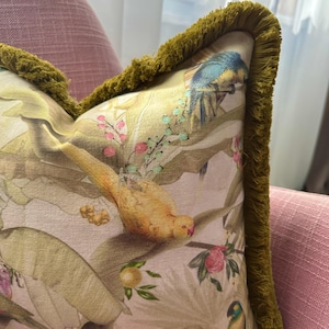 May include: A square throw cushion featuring a botanical and bird print on a light background, decorated with a thick olive green fringe trim along the edges. The fabric displays illustrations of yellow and blue birds, green leaves, and small pink berries.