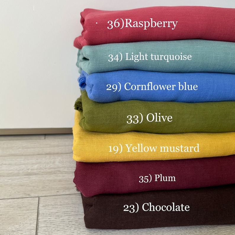 May include: A stack of folded fabric pieces in various colors, labeled with numbers and names: 36) Raspberry, 34) Light turquoise, 29) Cornflower blue, 33) Olive, 19) Yellow mustard, 35) Plum, and 23) Chocolate. The textiles have a visible woven texture.