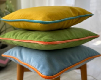 May include: Three decorative throw pillows stacked on a wooden stool. The top pillow is yellow with a teal trim, the middle pillow is green with an orange trim, and the bottom pillow is blue with an orange trim.