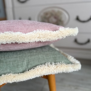 Peut inclure: Deux coussins décoratifs avec des bords frangés. Le coussin du haut est de couleur rose poussiéreux et le coussin du bas est vert mat. Les coussins sont posés sur un tabouret en bois.