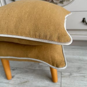 Könnte beinhalten: Zwei senfgelbe Kissen mit weißen Paspeln. Die Kissen liegen übereinander und befinden sich auf einem Holzhocker.