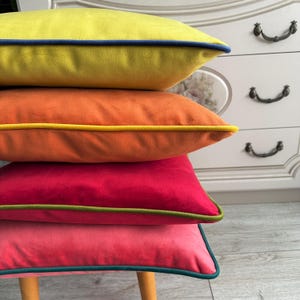 Puede incluir: Cuatro cojines cuadrados de colores apilados. El cojín superior es amarillo con ribete azul marino, seguido de naranja con ribete amarillo, rojo con ribete verde y rosa con ribete turquesa. Los cojines están sobre un taburete de madera.