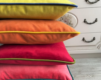 May include: Four colorful square throw pillows stacked on top of each other. The top pillow is yellow with a navy blue trim, followed by orange with yellow trim, red with green trim, and pink with teal trim. The pillows are on a wooden stool.