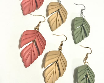 May include: Earrings in various colors, including dusty rose, beige, and sage green, each featuring a leaf design. The earrings have metal hooks, some gold-toned and some silver-toned, and are arranged on a white background.