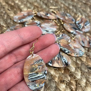 May include: A pair of oval-shaped earrings with a white, black, and gold abstract design. The earrings are made of resin and have gold-toned hooks.