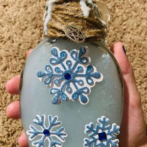 May include: A glass bottle with a blue liquid and snowflake decorations. The bottle is wrapped in twine and has a white wax seal.