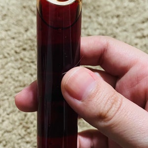 May include: A small glass bottle with a cork stopper and a wooden pentagram charm hanging from the top. The bottle is filled with a dark red liquid.