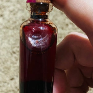 May include: A small glass bottle with a dark red liquid inside. The bottle has a gold-colored cap with a pink top.