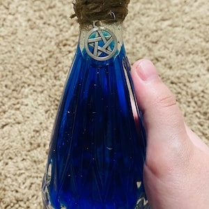 May include: A blue glass bottle with a pentagram charm and a twine-wrapped stopper. The bottle has a textured, faceted design.