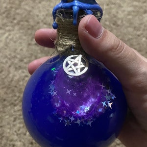 May include: A blue glass bottle with a purple and silver glitter interior. The bottle has a blue wax seal and a silver pentagram charm.