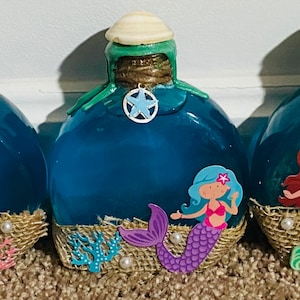 May include: Three teal blue glass bottles with mermaid themed decorations. Each bottle has a burlap and twine base with a seashell, coral, and pearl accents. The bottles are filled with a blue liquid and have a starfish charm hanging from the top.