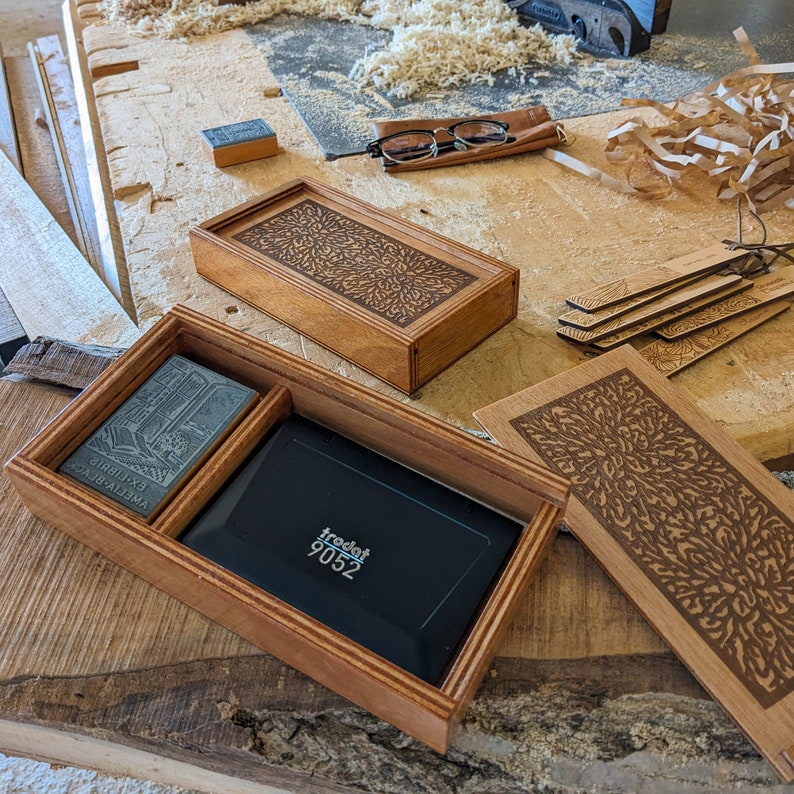 Two boxes are displayed: one with its lid removed, featuring an Ex Libris stamp and its accompanying stamp pad, with the other box sitting directly behind it, with its lid closed. In the foreground are tools, a pair of glasses, a stamp and sawdust.