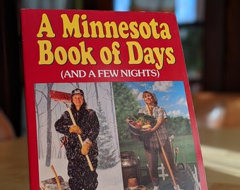 May include: A red book titled "A Minnesota Book of Days (And a Few Nights)" by Howard Mohr. The cover features a split image: a person in winter gear and a person holding vegetables. The book is displayed on a gold-colored stand.