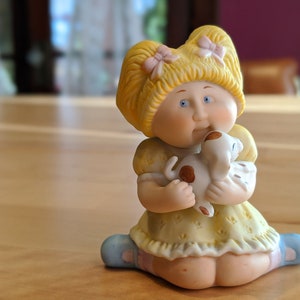 Pode incluir: Estatueta de cerâmica de uma boneca loira com olhos azuis, vestida com um vestido amarelo e segurando um cachorrinho branco com manchas marrons. A boneca tem laços cor-de-rosa no cabelo e está ajoelhada.