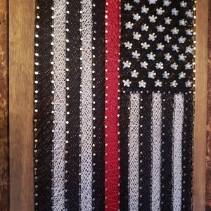 May include: A string art piece depicting the American flag with a red stripe in the center. The flag is made with white and black string wrapped around nails hammered into a wooden board.