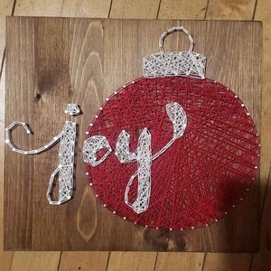 以下が含まれることがあります： 白い糸を釘に巻きつけた「喜び」という言葉が書かれた木製の看板。赤いクリスマスオーナメントも白い糸で飾られています。