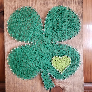 Peut inclure: Art de fil vert représentant un trèfle à quatre feuilles avec un petit cœur jaune au centre.