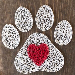 Può includere: Un'impronta di zampa realizzata a mano con string art e un cuore rosso al centro. L'impronta è realizzata con filo bianco e chiodi, con il cuore in filo rosso. L'opera d'arte è esposta su uno sfondo di legno.