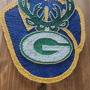 Puede incluir: Arte de hilo con el logotipo de los Green Bay Packers con una "G" verde y blanca rodeada de un contorno amarillo y azul y una cabeza de ciervo verde con cuernos en la parte superior.