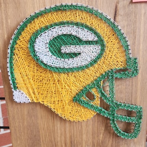 May include: Green Bay Packers string art featuring a football helmet with a green and white 'G' on the side. The art is made with yellow, green, and white string wrapped around nails hammered into a wooden board.