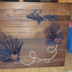 May include: A wooden sign with two outlines of the state of Michigan, each with a red heart in the center. The outlines are made of string wrapped around nails. A silver string connects the two hearts.