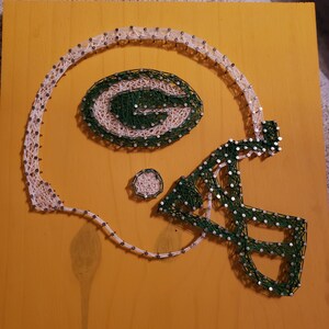 May include: Green Bay Packers football helmet string art on a yellow wooden background. The helmet is made of white and green string wrapped around nails.