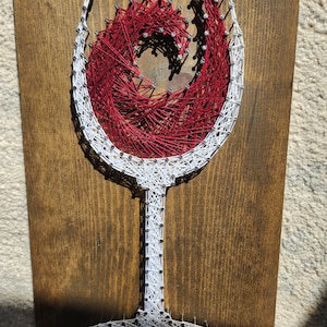 May include: A wine glass made with string art on a wooden background. The glass is outlined in white string and filled with red string in a spiral pattern.