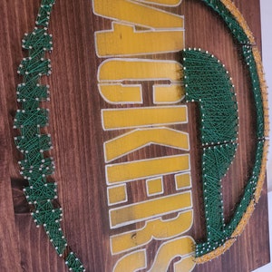 May include: Green and gold string art of the Green Bay Packers logo on a wooden background.
