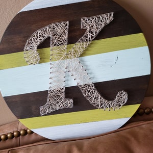 May include: A round wooden sign with a white, brown, and yellow striped background. The letter K is created using string art on the sign.