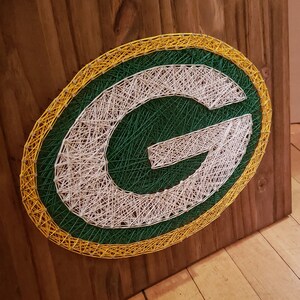 May include: Green Bay Packers logo string art on a wooden background. The logo is made of white and green string wrapped around nails. The logo is surrounded by a yellow string.