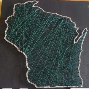 Può includere: Un'opera d'arte con filo raffigurante lo stato del Wisconsin. Il contorno è definito da chiodi argentati, con filo verde che si incrocia all'interno. L'opera è montata su uno sfondo nero, creando un contrasto sorprendente.