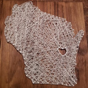 Può includere: Un'opera d'arte con filo raffigurante lo stato del Wisconsin, realizzata con filo bianco su sfondo in legno. Il design presenta un ritaglio a forma di cuore. È un oggetto decorativo.