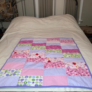 Puede incluir: Una colcha de retazos con un borde morado y una variedad de cuadrados estampados en rosa, verde y blanco.