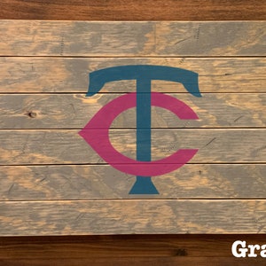 K&ouml;nnte beinhalten: Ein Holzschild mit einem grauen, blauen und rosafarbenen Minnesota Twins Logo. Das Schild ist aus wiederverwendetem Holz gefertigt und hat einen rustikalen Look.