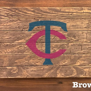 K&ouml;nnte beinhalten: Ein Holzschild mit einem stilisierten "TC"-Logo in Teal und Maroon. Das Schild ist aus wiederverwendetem Holz gefertigt und hat einen rustikalen, abgenutzten Look. Das Wort "Brown" ist unter dem Schild gedruckt.