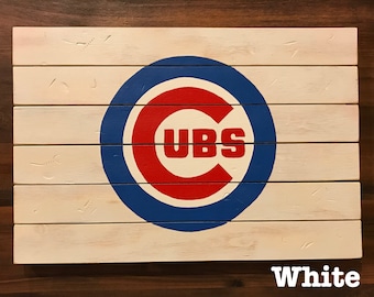 May include: White wooden sign with a blue and red Chicago Cubs logo. The logo is a circle with a "C" in the center and "UBS" written inside the "C".