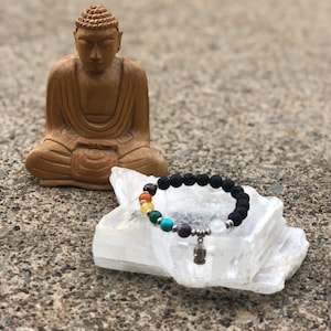 Puede incluir: Un brazalete de piedra de lava negra con un dije de plata de un Buda meditando. El brazalete presenta siete piedras de chakra en colores arcoíris. El brazalete descansa sobre un cristal blanco.