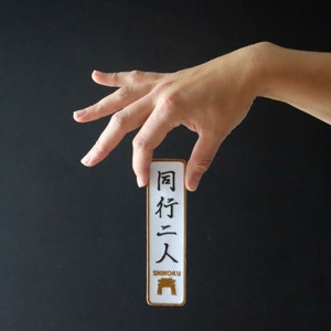 Könnte beinhalten: Ein weißer rechteckiger Patch mit schwarzen japanischen Schriftzeichen, die "同行二人" lauten, und dem Wort "SHIKOKU" in goldenen Buchstaben darunter. Unter dem Wort "SHIKOKU" befindet sich ein goldenes stilisiertes Bild eines Torii-Tores.