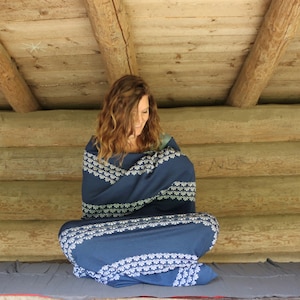 Può includere: Una persona seduta su una panca di legno, avvolta in una coperta a motivi blu e bianchi. La coperta ha un motivo geometrico bianco su uno sfondo blu scuro.