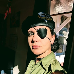 Puede incluir: Boina negra con un parche de "Diamond Dogs", usada con un parche en el ojo negro y una camisa verde. El parche en el ojo está cosido con hilo blanco. También se ve un collar de gargantilla negro.