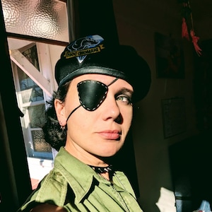 May include: Black beret with a leather patch featuring the text "DIAMOND DOGS" and a dog graphic. A black leather eye patch is worn over one eye. The person is wearing a green shirt and a black choker.