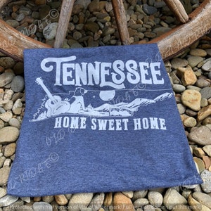 May include: A blue t-shirt with a white graphic design of the state of Tennessee with a guitar and a dog sitting on a mountain. The text "Tennessee" and "Home Sweet Home" are printed in white.