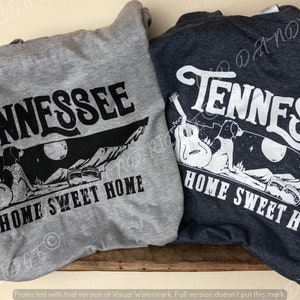 May include: Two t-shirts with a graphic design featuring the state of Tennessee with a silhouette of a couple sitting on a hill with a guitar and the words "Home Sweet Home". One t-shirt is grey and the other is dark blue.