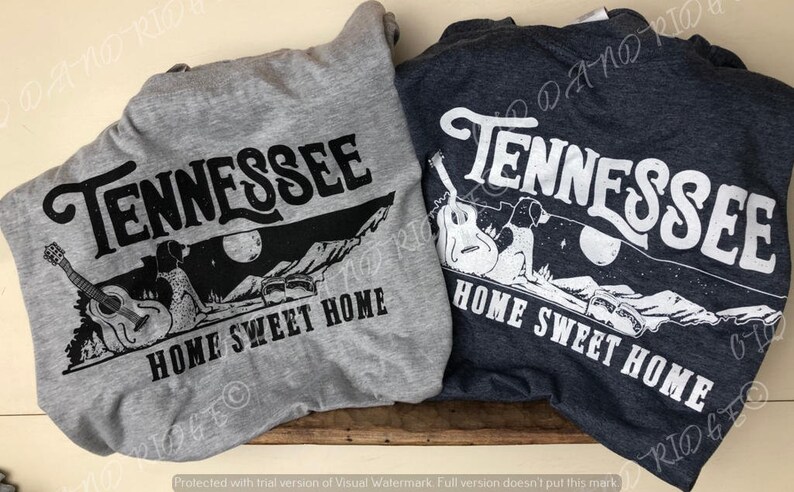 May include: Two t-shirts with a graphic design featuring the state of Tennessee with a silhouette of a couple sitting on a hill with a guitar and the words "Home Sweet Home". One t-shirt is grey and the other is dark blue.